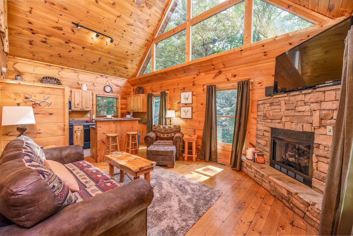 A wood-paneled living area features a cozy sofa facing a stone fireplace. Natural light floods the space through large A-frame windows, highlighting the warm wood tones and inviting decor. A kitchen area with wooden cabinetry and a bar top is visible in the background.