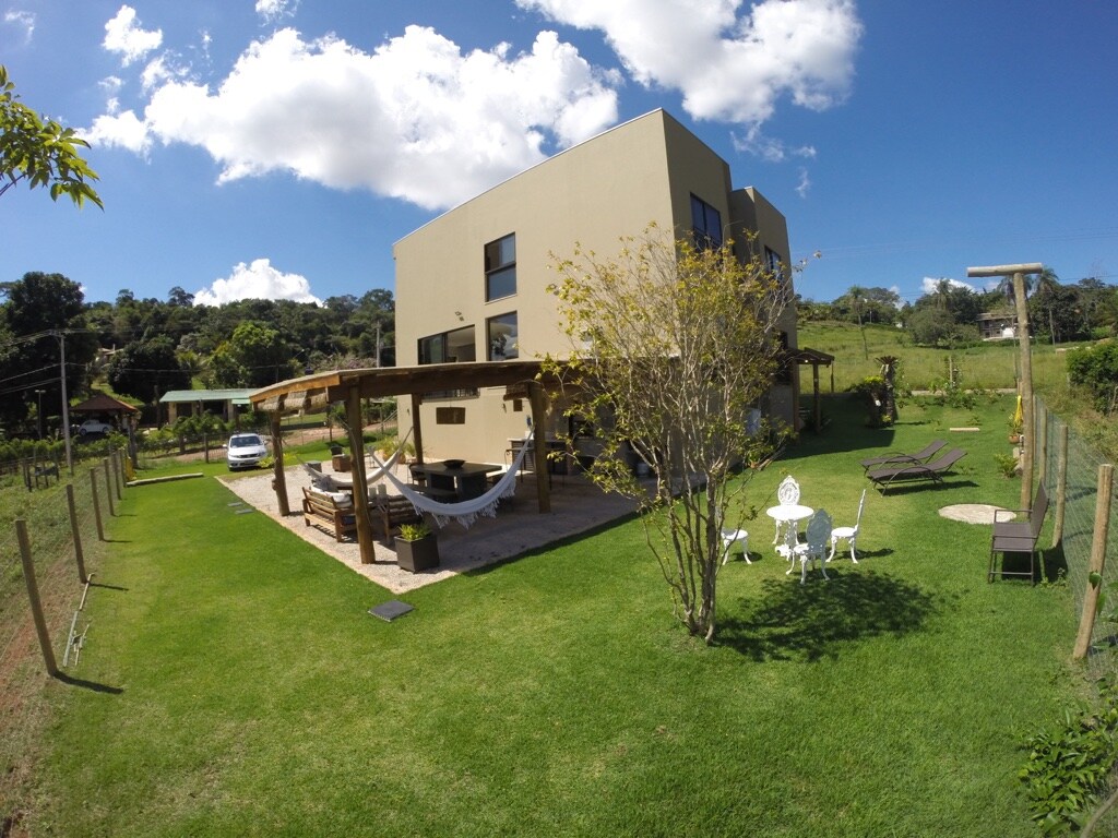 The exterior of the house is surrounded by a well-maintained grassy area, featuring a variety of outdoor seating options including lounge chairs and a dining set. A hammock is suspended under a wooden pergola, and a tree offers shade in the lush garden.