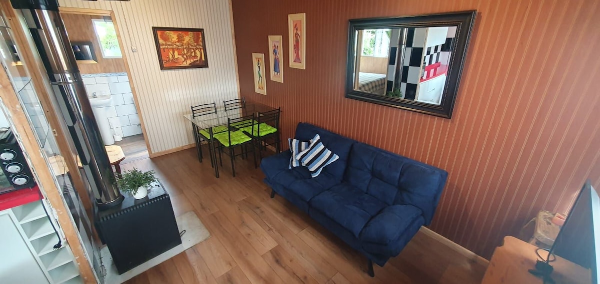 An inviting living space features a dark blue couch complemented by decorative pillows. A small dining table with green accents and black chairs is positioned nearby. A large mirror reflects the room's warmth, while wooden flooring adds to the cozy ambiance.