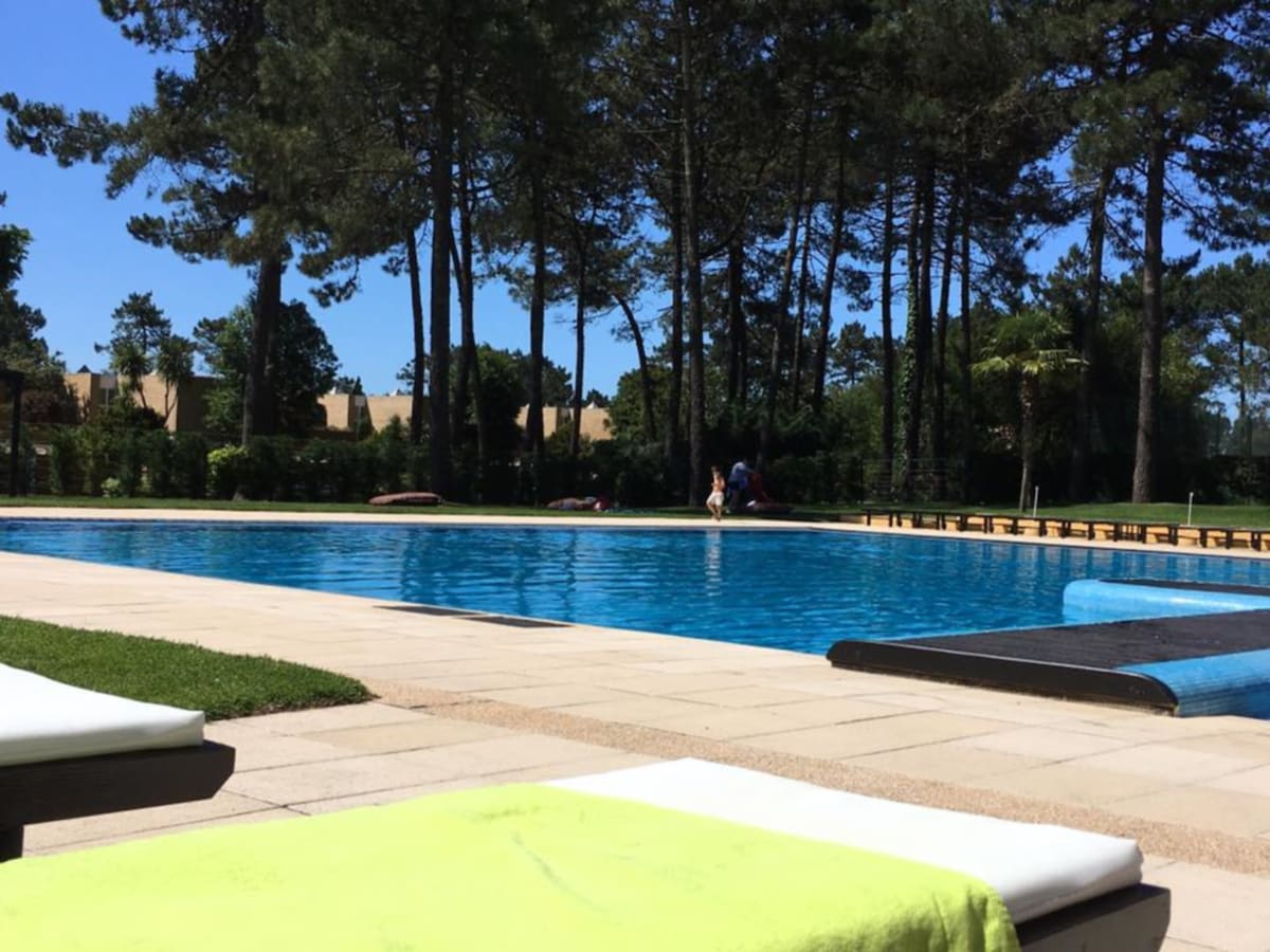 A spacious pool area is surrounded by tall trees, providing a natural setting. The clear blue water of the pool reflects bright sunlight. Poolside loungers are positioned on the paved deck, inviting relaxation. Lush greenery creates a serene backdrop in the distance.