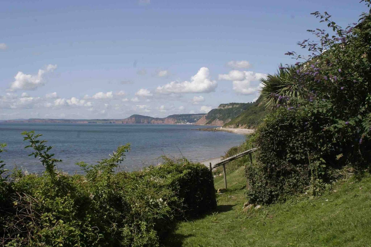 The image features a coastal view showcasing rolling hills and cliffs in the background, with a calm sea reflecting the sky. Lush greenery lines the foreground, leading towards the water's edge, enhancing the natural, serene atmosphere of the area.