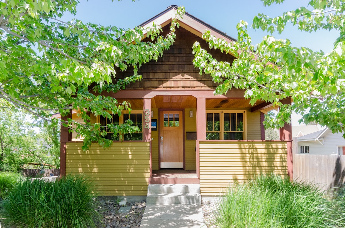 A historic bungalow exterior is highlighted by a blend of wood siding and a covered porch. Lush green foliage frames the entrance, showcasing the mature maple trees. Natural stone and native grasses create a welcoming approach to the front door, which is surrounded by large windows.