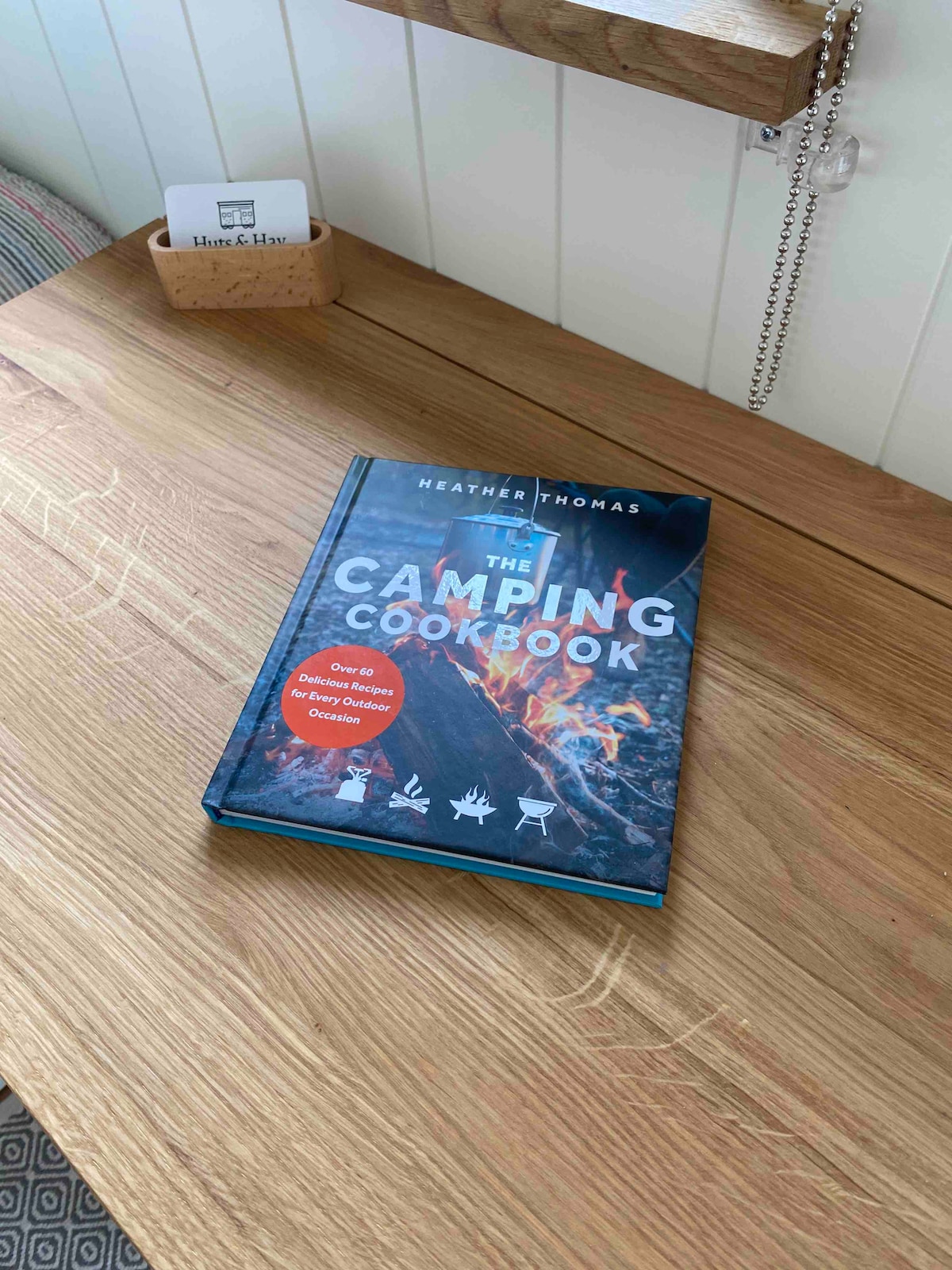 A camping cookbook is placed open on a wooden surface, showcasing its vibrant cover featuring a campfire scene. The book, titled 'The Camping Cookbook' by Heather Thomas, is accompanied by a small card holder on the table that adds to the rustic charm.