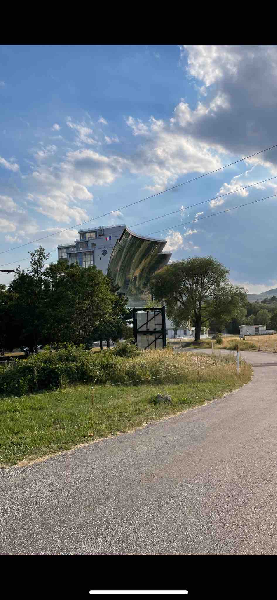 A unique structure is visible from a path lined with lush greenery. The building's distinctive design is complemented by a backdrop of trees and a partly cloudy sky, creating an inviting atmosphere for guests approaching the property.