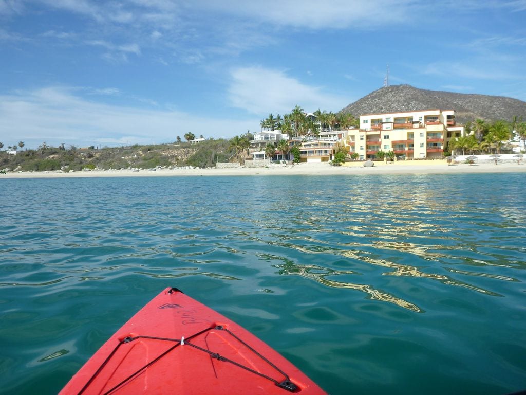 A red kayak is seen from the water, with a clear view of a sandy beach and a beachfront building in the background. Lush greenery and palm trees frame the area, while a mountain rises in the distance under a bright blue sky with scattered clouds.