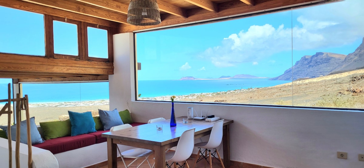 A light-filled living area features a large window offering expansive views of the sea and coastline. A wooden dining table is surrounded by modern chairs, while a cozy seating area includes cushions in earthy tones. Decorative items add a subtle touch to the space.