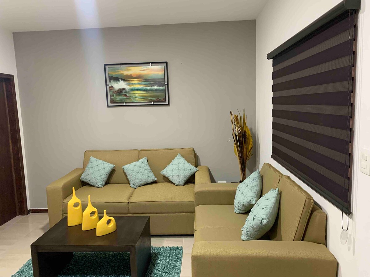 A comfortable seating area is presented with two sofas adorned with turquoise accent pillows. A black coffee table sits centrally on a textured blue rug, complemented by decorative yellow vases. A landscape painting and a modern window covering enhance the space.