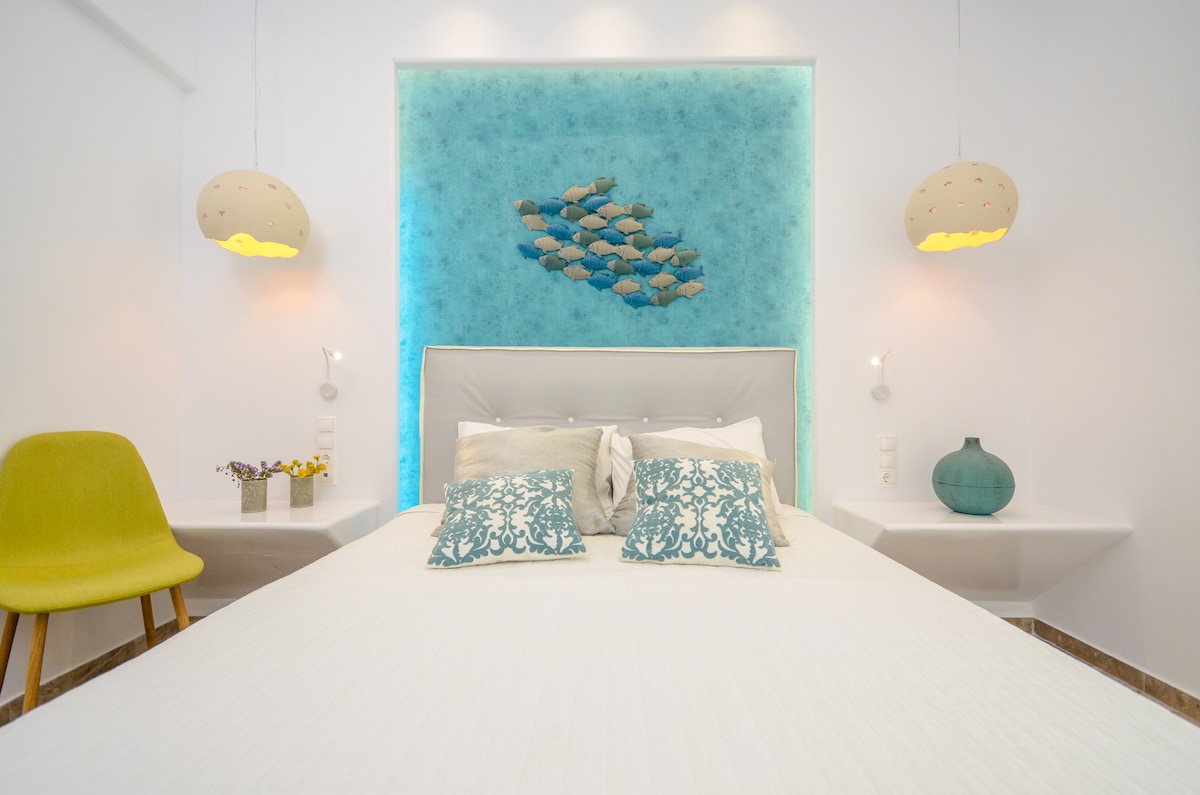A spacious bedroom features a light-coloured bed with decorative pillows and a unique fish wall art piece above the headboard. Soft lighting is provided by two hanging lamps, and a green accent chair is positioned beside the bed. Side tables with small decor items are visible.