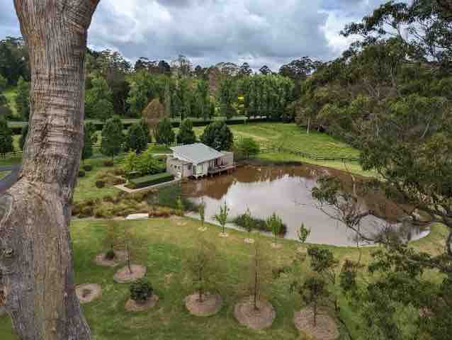 A quaint house is set beside a shimmering pond, surrounded by lush greenery and trees. The property features a well-maintained garden, including small shrubs and open grassy areas, creating a peaceful natural environment.