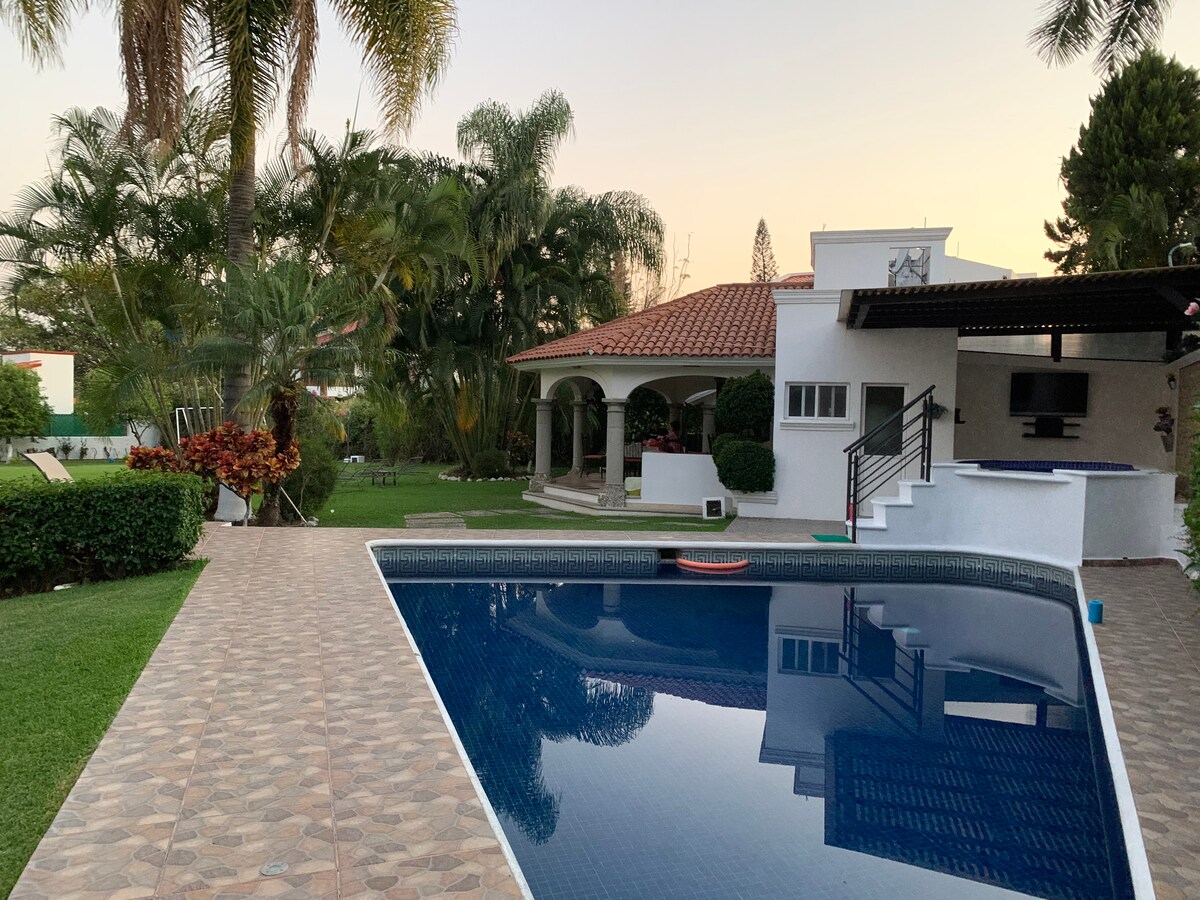 The outdoor area features a spacious swimming pool reflecting the surrounding landscape, complemented by well-manicured gardens and palm trees. A nearby palapa provides shaded seating, while the house showcases a blend of modern architecture and natural elements.