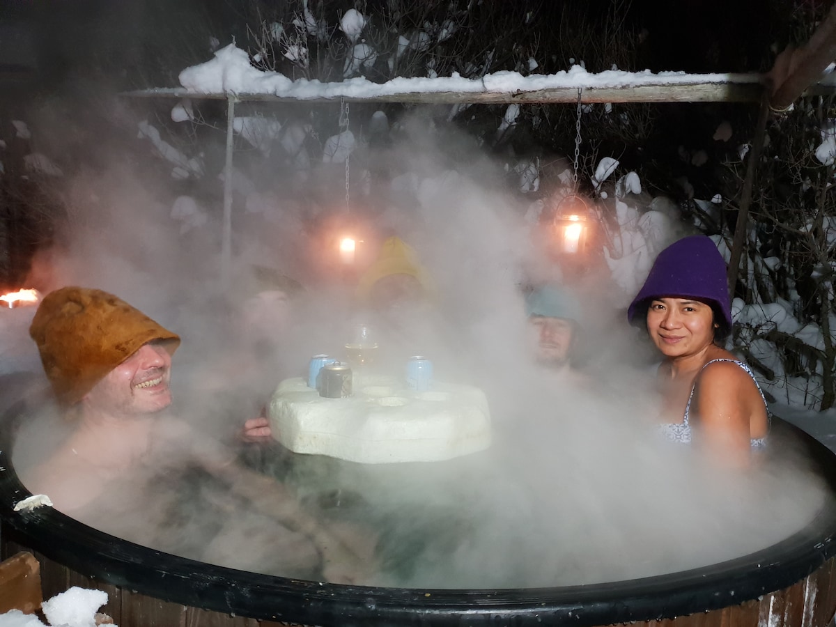 A round hot tub is surrounded by snow-covered landscape, with steam rising gently. Soft lighting hangs above, creating a serene ambiance. Several individuals are seated in the tub, enjoying the warm waters amidst the winter scenery.