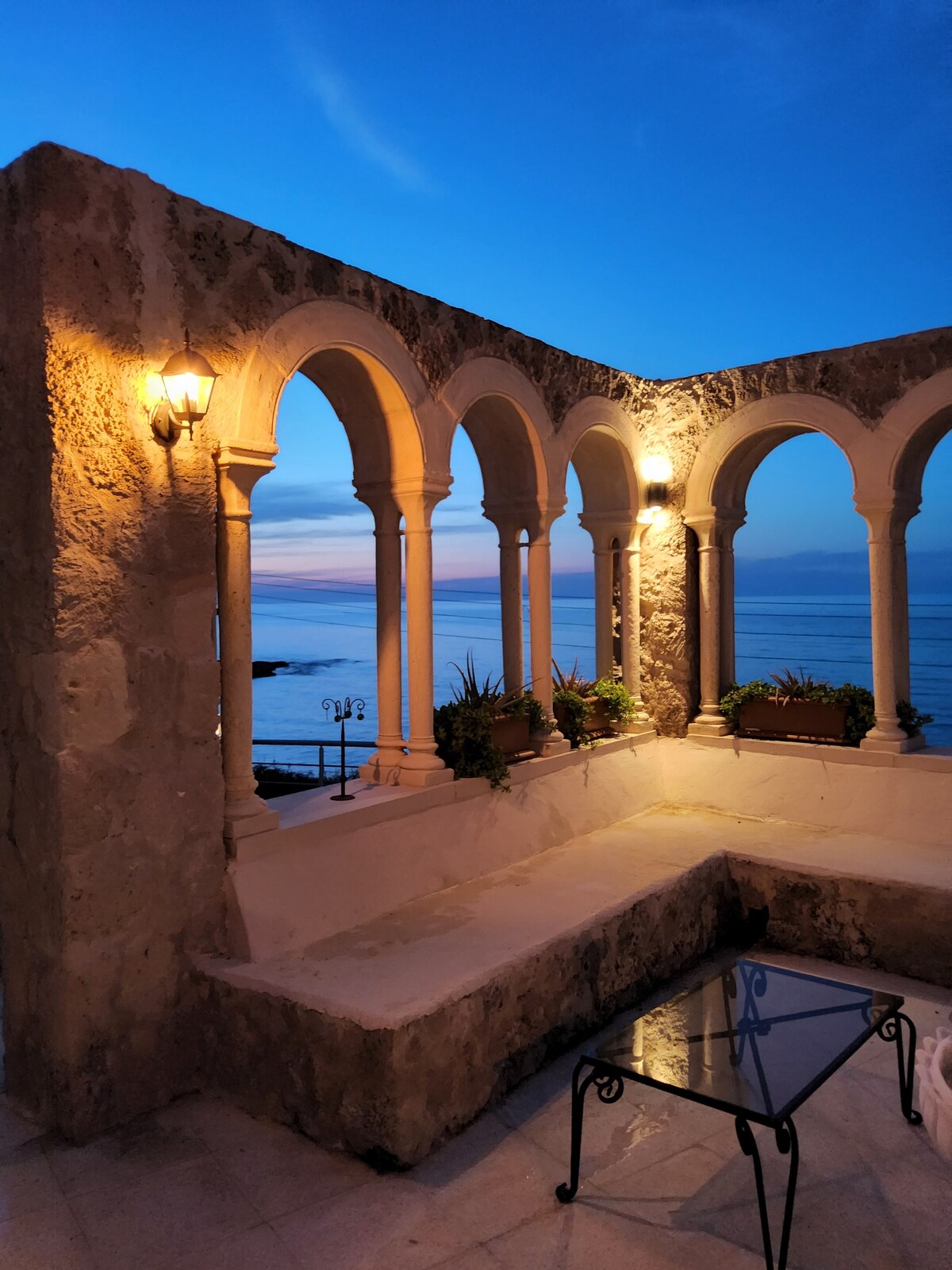 An open archway terrace offers views of the serene sea at dusk. Soft lighting illuminates the stone walls, accentuating the graceful arches. A glass-top table is centered on the seating area, surrounded by planters with greenery, creating a peaceful ambiance.