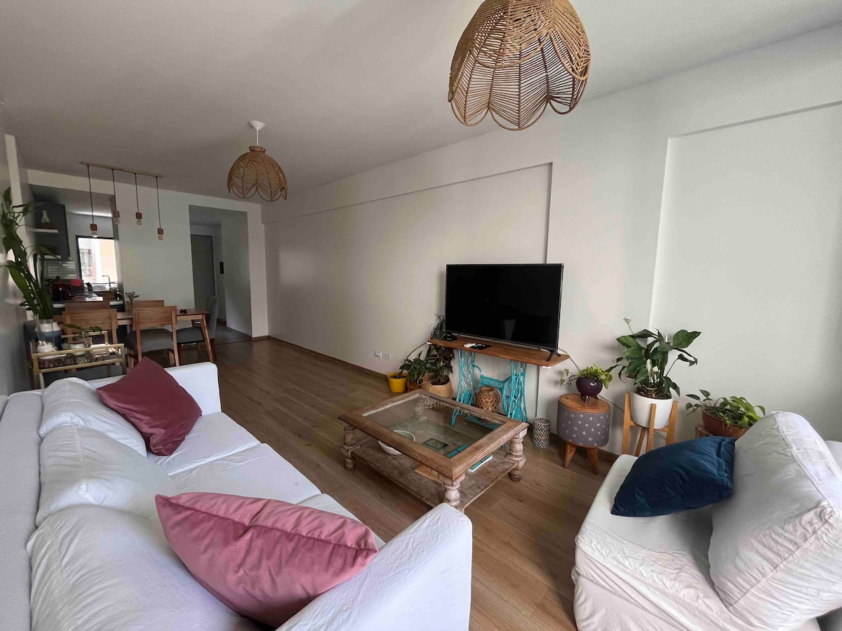 A spacious living room is showcased, featuring a comfortable white sofa adorned with colorful cushions. A wooden coffee table is centered in the area, while a television is positioned on a stand against the wall. Plants are arranged in the corner, enhancing the inviting atmosphere.