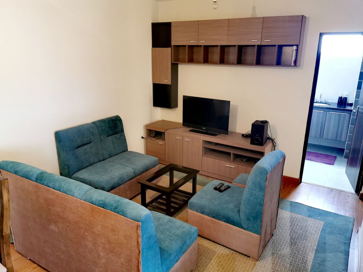 A modern living area is depicted, featuring a comfortable arrangement of teal sofas positioned around a glass coffee table. A television is mounted on a wooden media unit, with shelving displayed above. A doorway leads toward an adjoining kitchen area in the background.