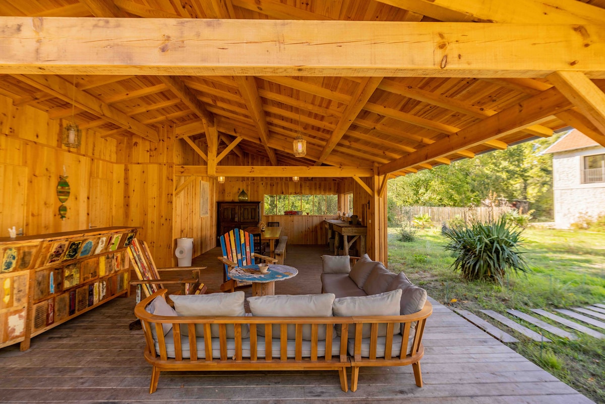 An outdoor living space features a wooden frame and ceiling, complemented by a spacious seating area with cushioned sofas. A table sits in the center, while bookshelves are positioned against the walls, adding an element of comfort and utility to the natural environment.