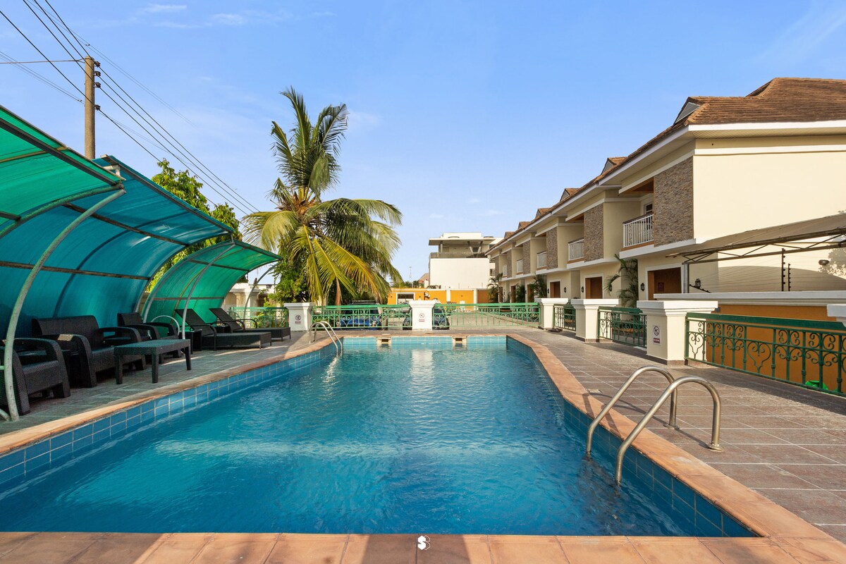 A spacious swimming pool is framed by lounge chairs under teal canopies. Surrounding greenery includes a palm tree, and a two-story building is visible in the background, showcasing a well-kept exterior.