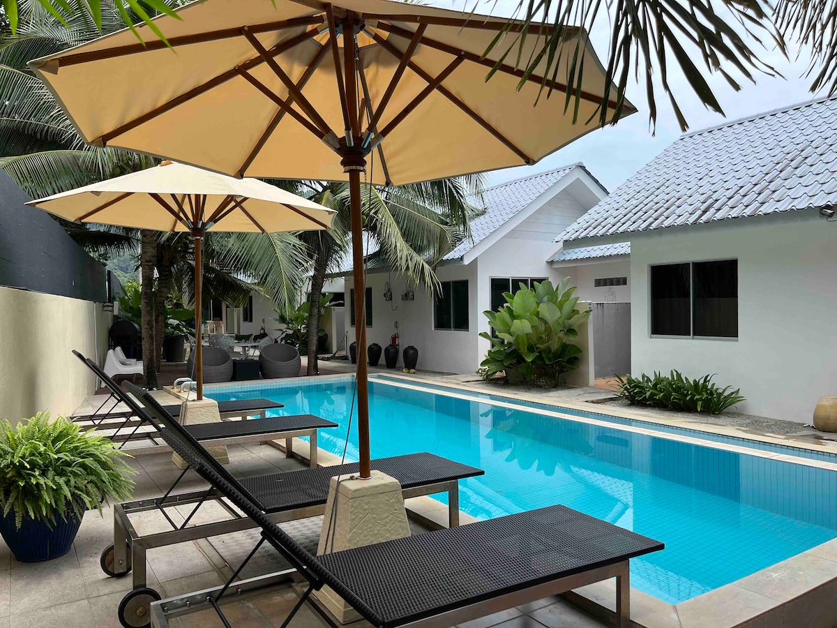 A swimming pool is surrounded by loungers and umbrellas, offering a spot for relaxation. Tropical foliage and the villa's white exterior are visible in the background, contributing to a serene atmosphere, while the gentle slope of the pool adds to its inviting shape.