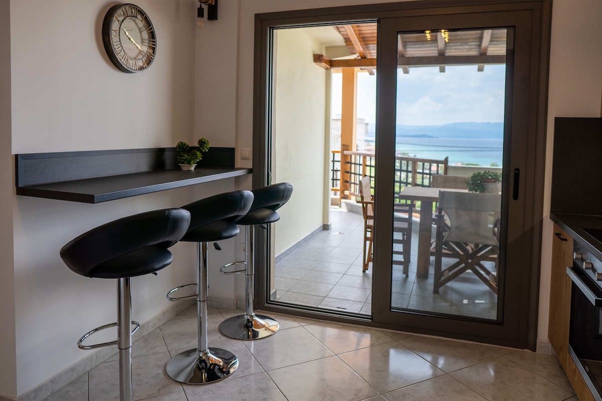 A modern kitchenette area features a sleek bar with three high-backed stools. A large sliding glass door opens to a balcony with views of the ocean, while a clock adorns the wall, adding to the contemporary design.