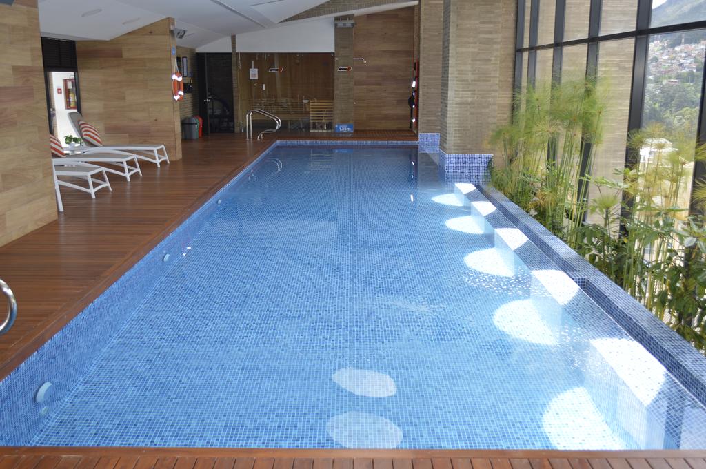 An indoor swimming pool is surrounded by wooden decking and large glass windows, providing ample natural light. Lounge chairs are placed along one side, and tropical plants are positioned at the edge, enhancing the tranquil setting.