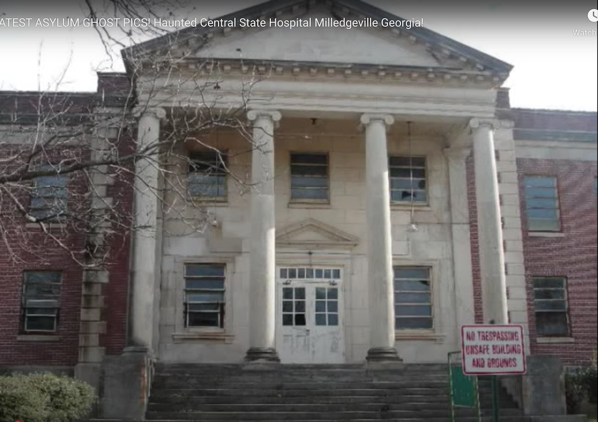 The historic facade of the Central State Hospital features towering columns and a broad entryway. The weathered brick exterior shows signs of aging, while a no trespassing sign is prominently displayed on the lawn, highlighting the restricted access to the buildings.