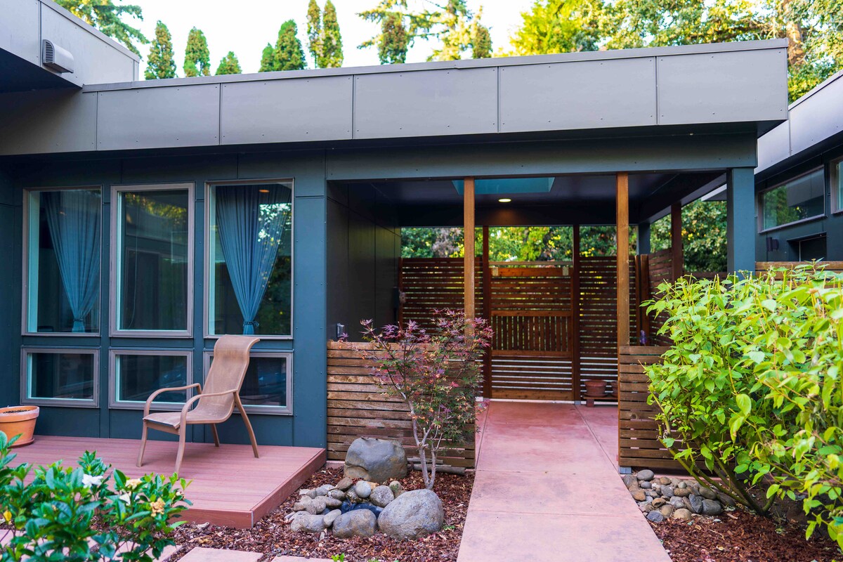 The entrance showcases a modern porch with large windows and inviting blue curtains. A comfortable chair is positioned on a wooden deck, surrounded by lush greenery and decorative rocks. A slatted wooden screen adds to the privacy of the entryway.