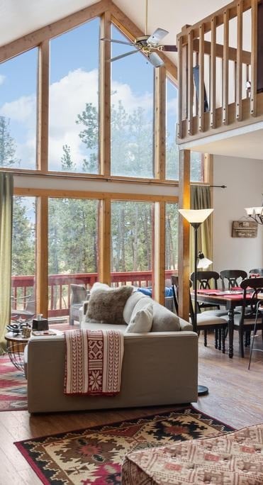 A spacious great room is bathed in natural light, featuring tall windows and a ceiling fan. A comfortable couch is positioned near a decorative rug, while a large dining area can be seen in the background, surrounded by forest views.