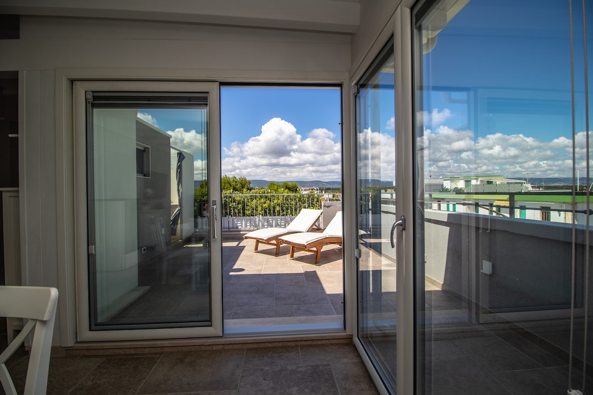 Large sliding glass doors provide access to a private terrace featuring sun loungers. The outdoor space offers views of the surrounding area, with bright blue skies and scattered clouds visible through the transparent panels. Natural light fills the interior space, highlighting the clean lines and modern design.