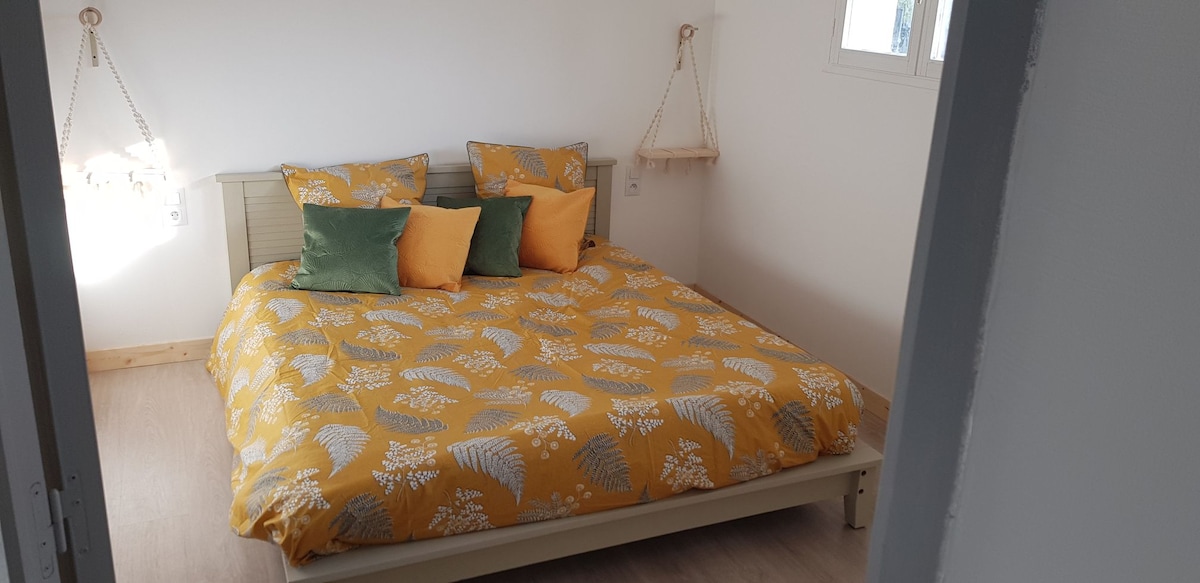 A bright sleeping area features a spacious bed with a yellow and gray patterned duvet, accented by several decorative pillows in green and orange. Natural light enters through a nearby window, creating a serene setting.