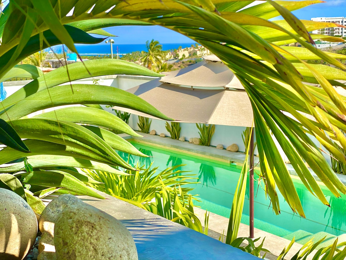 A serene swimming pool is framed by lush green foliage, with gentle waves reflecting sunlight. Sunshade structures are visible nearby, surrounded by landscaped areas. The distant ocean can be seen, enhancing the tranquil outdoor environment.