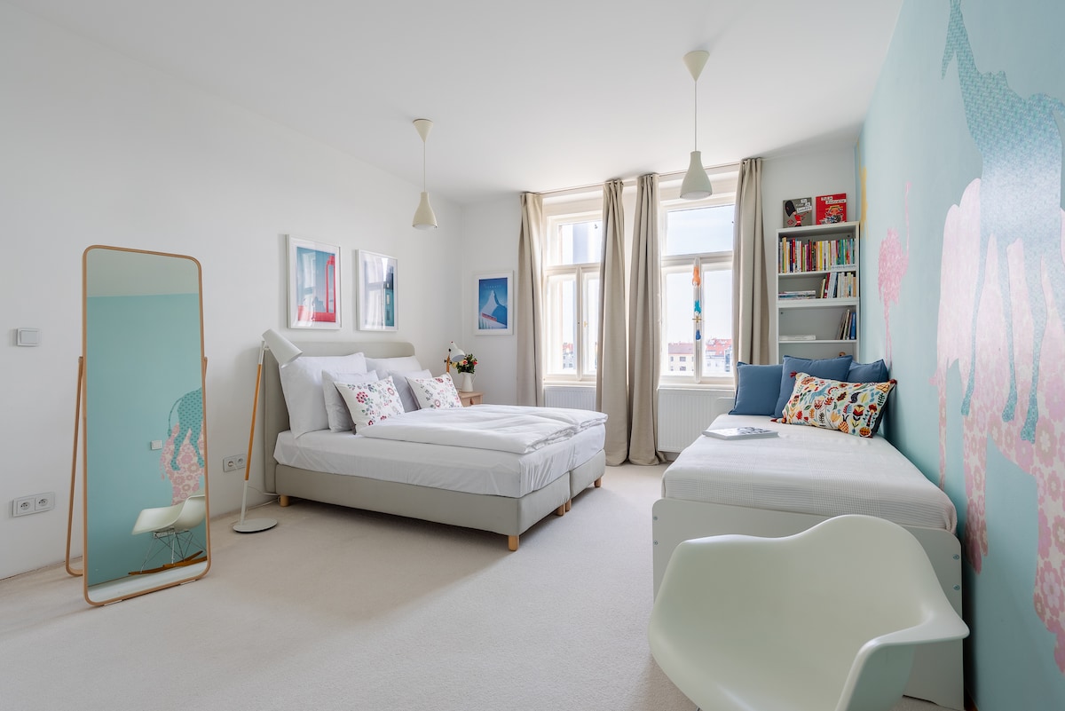 A bright bedroom features a spacious double bed and a single bed, positioned against a light blue wall adorned with artistic designs. Natural light fills the room through large windows dressed in light curtains. A full-length mirror stands beside the beds, and a bookshelf with decor is also present.