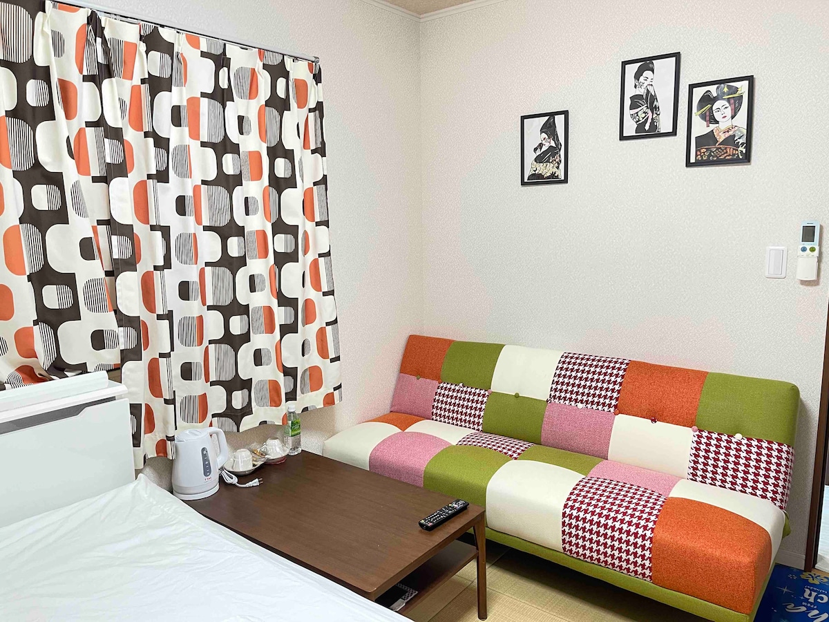 A colorful seating area is featured, showcasing a patchwork-style sofa in shades of green, orange, and white. A small wooden table is positioned in front, with a kettle and bottled water placed nearby. Playful patterned curtains add a lively touch to the room.