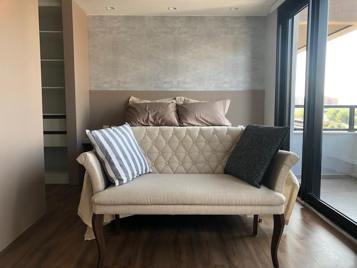 A modern seating area features a light-colored couch with tufted detailing and cozy, contrasting pillows. Natural light streams in from large glass doors, creating an airy atmosphere. A soft, neutral wall complements the floor, enhancing the overall simplicity of the space.