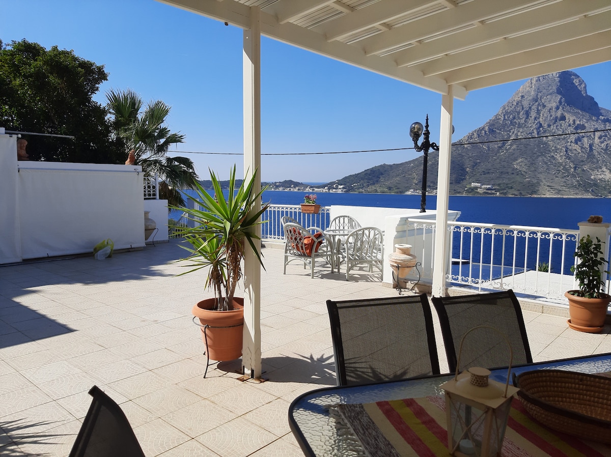 A spacious terrace offers panoramic views of the sea and surrounding mountains. Outdoor seating is arranged for relaxation, with potted plants adding a touch of greenery. Sunlight brightens the area, reflecting off the tiled floor, creating an inviting spot for outdoor dining.