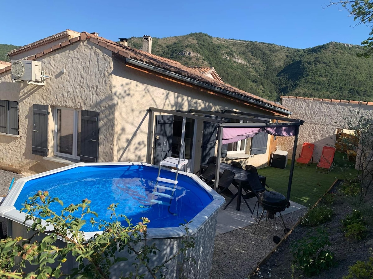 A private outdoor space features a round pool surrounded by sun loungers and a shaded seating area under a canopy. The property is located against a backdrop of green hills, with the sky visible in clear blue. Nearby, an outdoor barbecue setup is present.