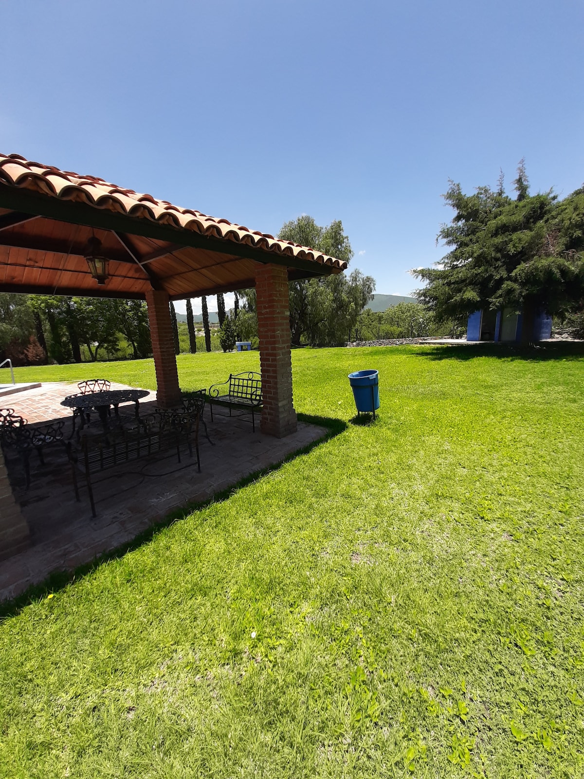 A shaded palapa is featured with a tiled floor and several metal chairs surrounding a central table. Lush green grass extends into the background, where trees are visible. A blue trash bin is placed nearby, contributing to the neat appearance of the outdoor area.