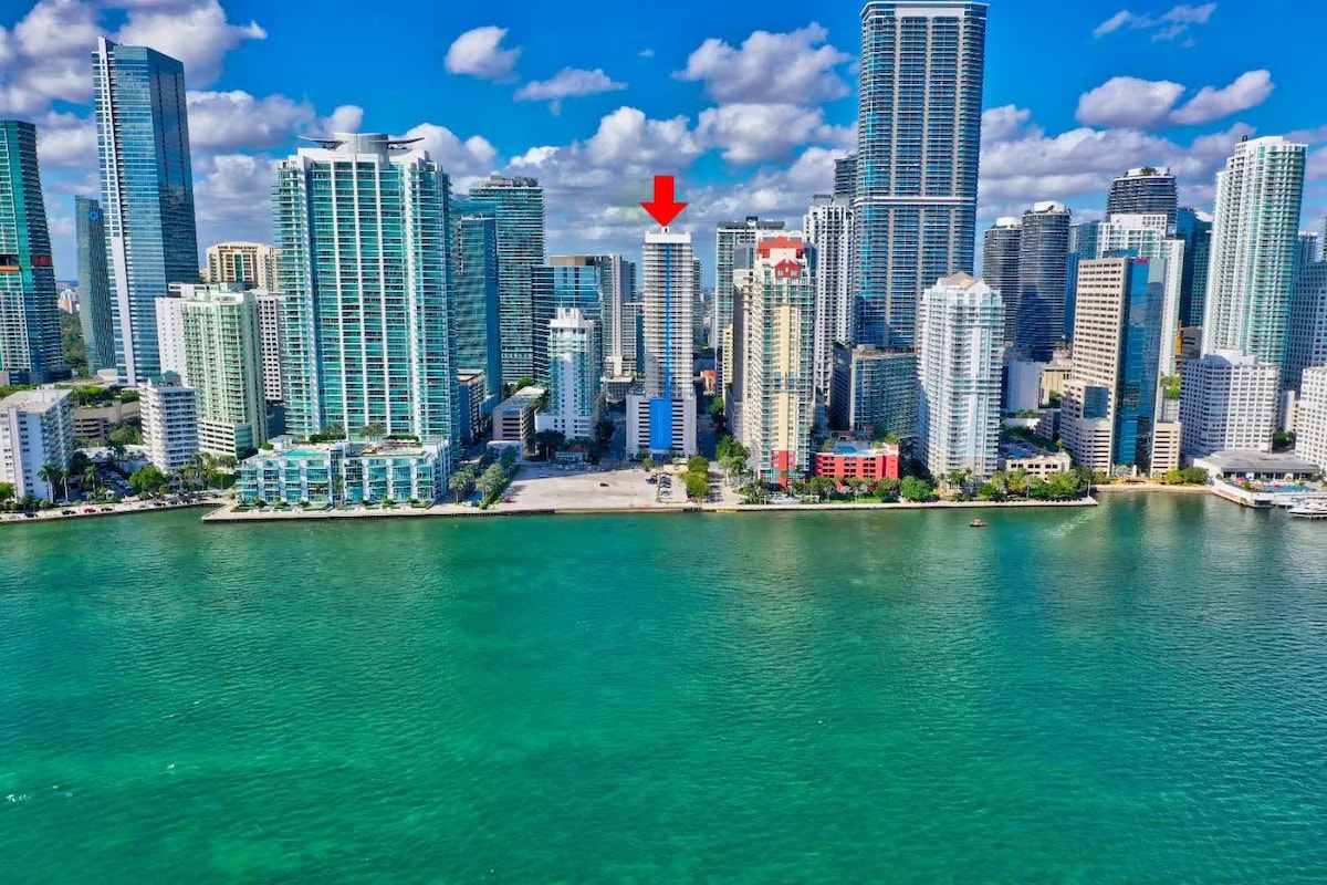 The city skyline of Miami is prominently displayed, with various high-rise buildings reflecting in the turquoise waters of the bay. A red arrow points to the condo, indicating its location within this vibrant urban landscape. Fluffy white clouds dot the blue sky, enhancing the scene.
