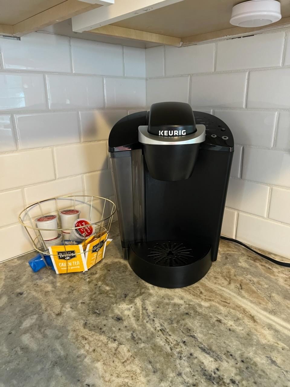 A black Keurig coffee maker is positioned on a granite countertop beneath overhead cabinetry. A wire basket holds various coffee pod flavors, including a selection of tea, positioned nearby. The clean white tile backsplash complements the modern kitchen design.