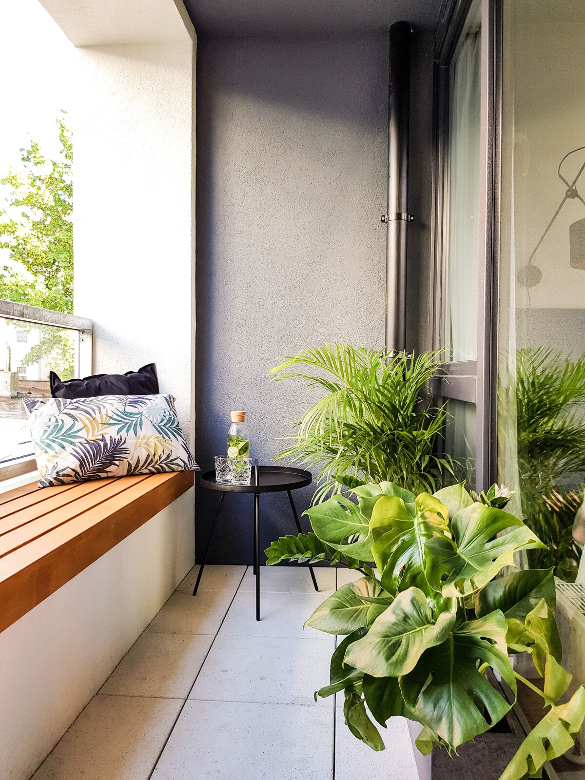 A cozy loggia features a wooden bench accented by a decorative pillow, providing comfortable seating. A small round table is placed beside a collection of lush green plants, creating a serene and inviting outdoor space. Large windows offer a view of the surroundings.