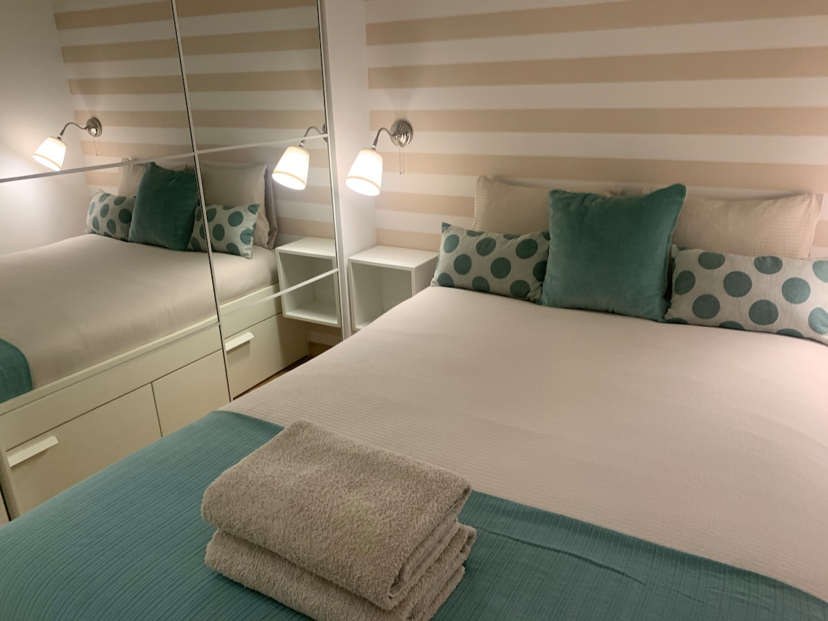 A comfortable bedroom is presented with a spacious bed adorned with soft linens and decorative pillows. The room features a mirrored wardrobe, enhancing the sense of space, while warm lighting from wall sconces creates a cozy ambiance. Clean towels are neatly arranged on the bed.