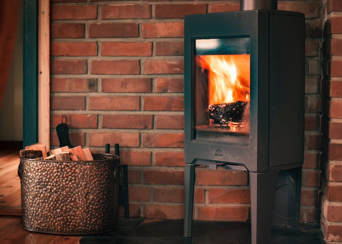 A wood-burning stove is positioned against a wall of warm red bricks, radiating inviting warmth. Firewood is stacked neatly in a woven basket nearby, enhancing the cozy atmosphere of the space. The stove features a clear glass front, showcasing the flickering flames.