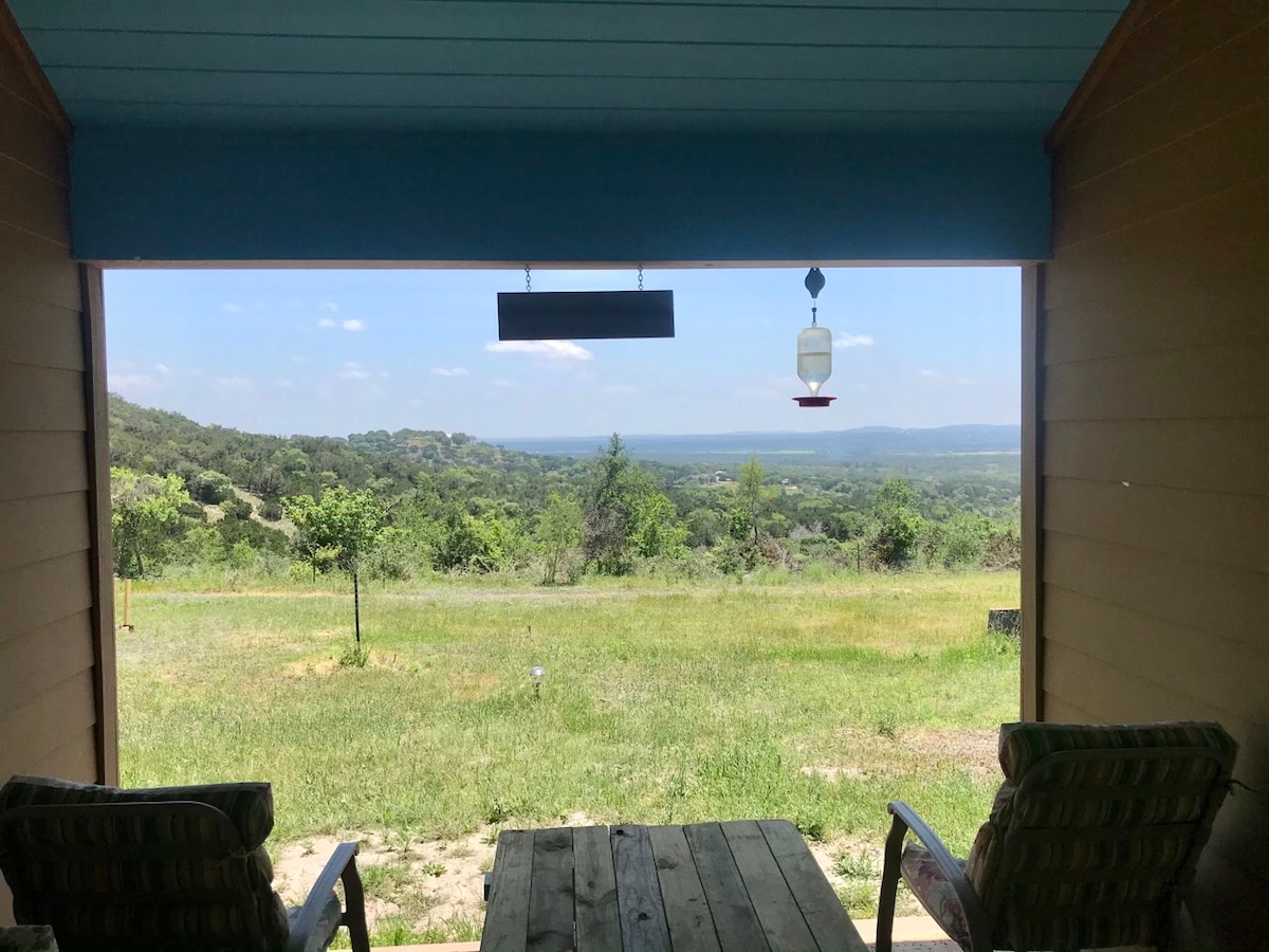 A serene view is captured from a covered porch, featuring a wooden table and two chairs. Beyond, rolling hills and lush greenery extend across the landscape, with a bright blue sky adding to the expansive scene.