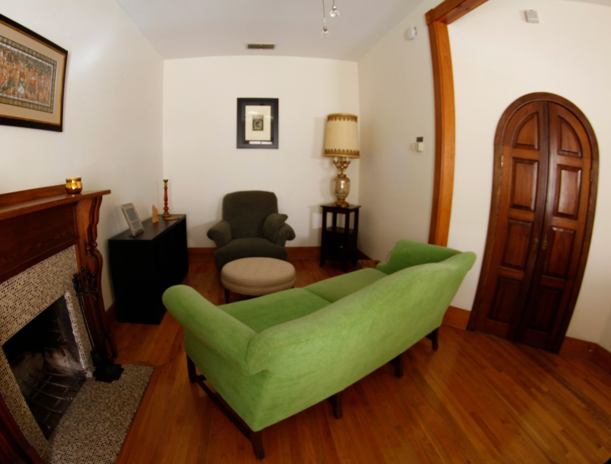 A cozy living area features a green sofa opposite a traditional fireplace framed by a wooden mantel. A round ottoman sits in the center, and an arched doorway offers an inviting entrance. A decorative lamp stands next to an accent table, adding to the room's charm.