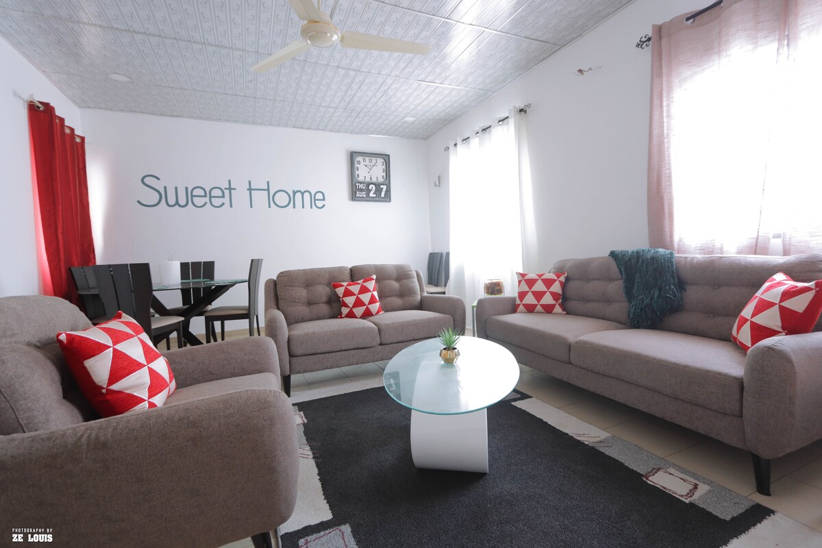 A welcoming living area is presented, featuring a round glass table centered among two gray sofas adorned with red and white cushions. A wall clock is visible above the seating, and a dining table can be noticed in the background, complemented by neutral-colored decor.