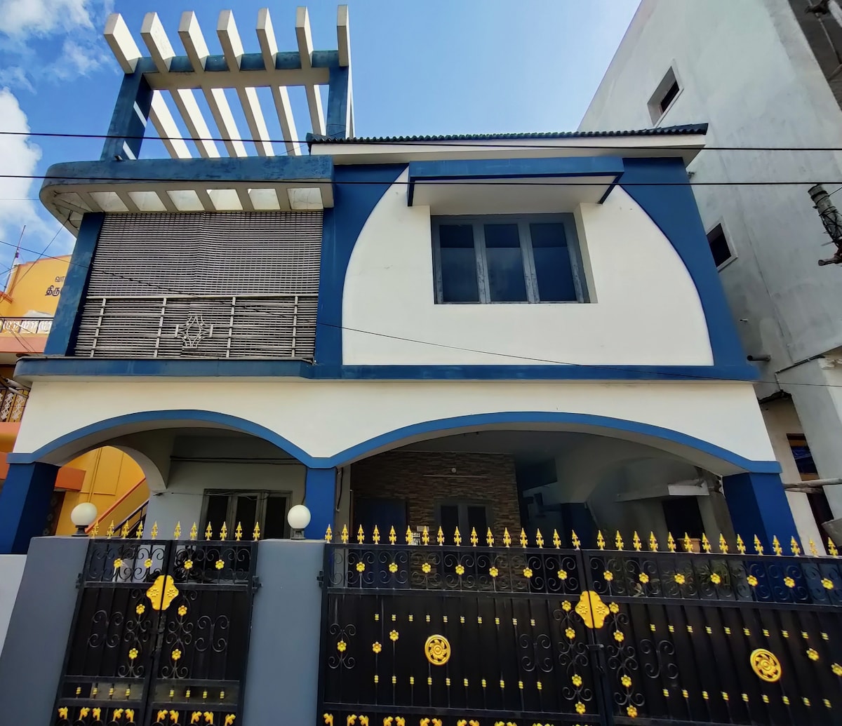 The exterior of the homestay showcases a two-story structure accented with blue and white walls. An arched entrance leads to the private entrance. A gated fence with decorative golden elements adds security. A pergola spans the upper level, highlighting the architectural design.