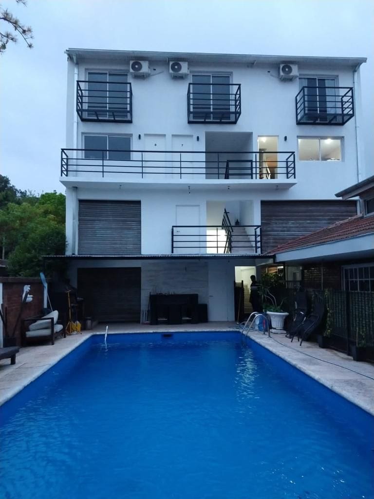 A multi-level building is presented with several balconies and large windows. A spacious pool is located in the foreground, featuring a clear blue surface. Poolside seating and lounging areas are visible, surrounded by greenery that adds to the space's serene environment.