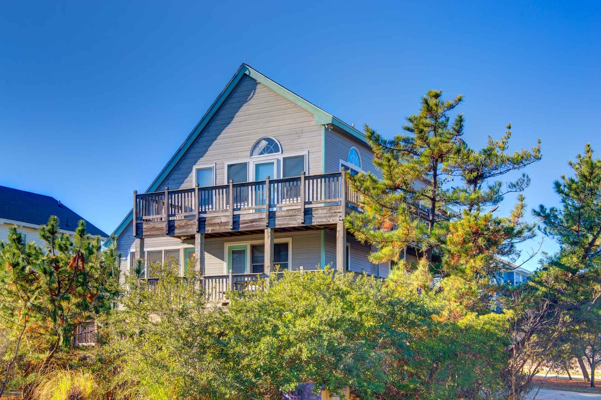 The exterior of the beach house features a two-story structure with a pitched roof, surrounded by greenery. A wrap-around deck is visible on the upper level, complemented by multiple windows that invite natural light. Tall pine trees and ornamental shrubs add lushness to the setting.