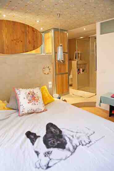 A cozy bedroom is highlighted by soft bedding adorned with an illustration of a dog. A spacious en-suite shower area is visible in the background, featuring elegant wood and glass elements. The ceiling showcases a unique design, complementing the overall inviting atmosphere.