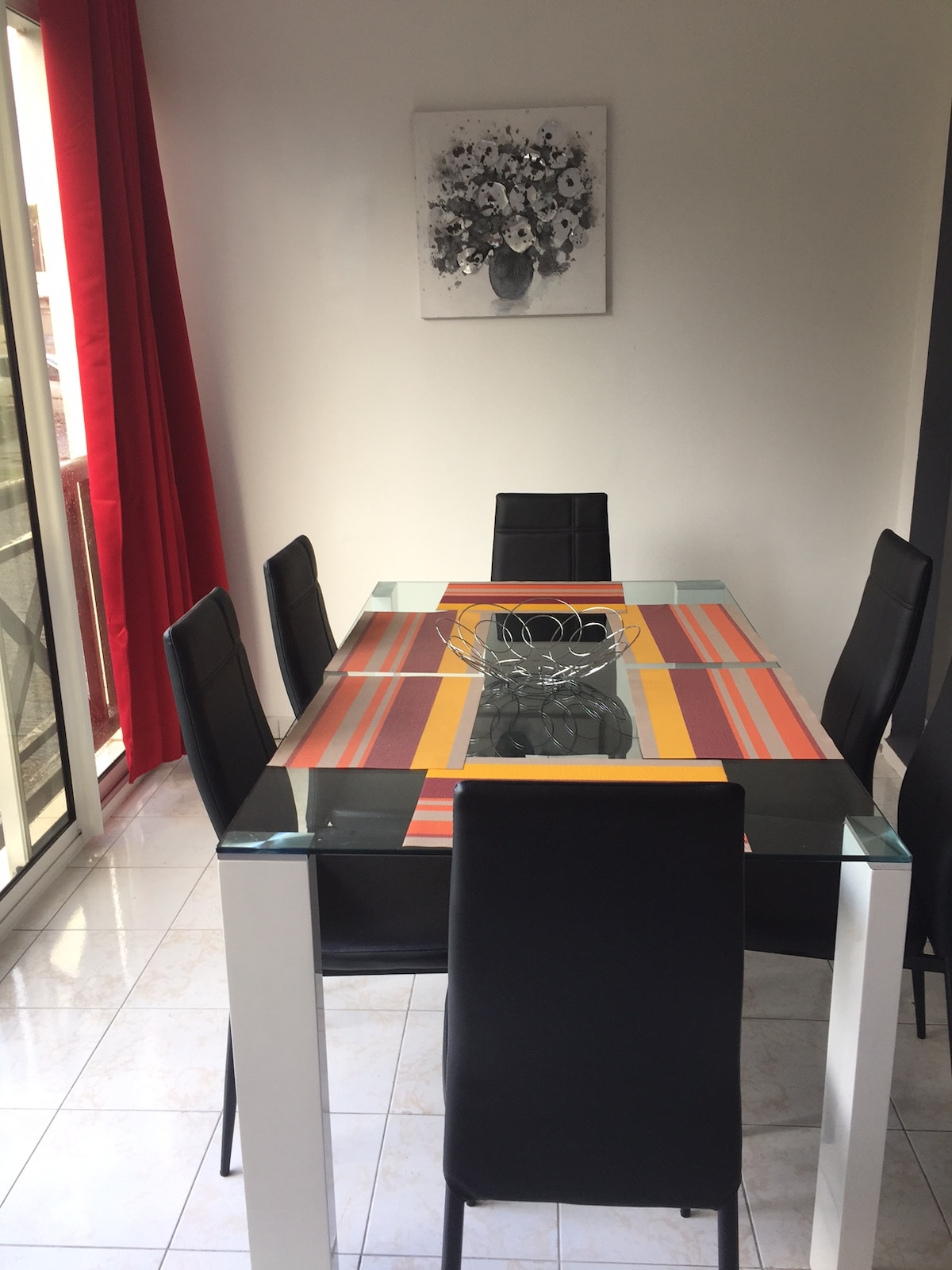 A dining area features a glass-top table, surrounded by six black chairs. A colorful striped table runner adds a vibrant touch, and a decorative bowl sits at the center. Natural light enters through the adjacent glass sliding doors, complemented by red curtains.