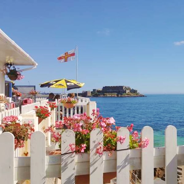 A white picket fence encloses a seaside area adorned with vibrant pink flowers. A sun umbrella provides shade, while a flag flutters in the breeze. In the background, the serene blue water reflects the sky, and a castle is visible on a distant island.