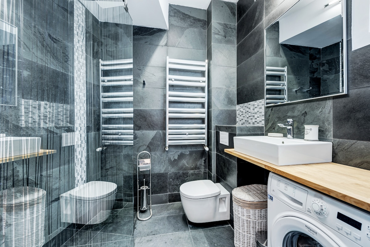 A modern bathroom is highlighted by sleek slate tiles and contemporary fixtures. A wall-mounted towel warmer and a stylish sink with a wooden counter are featured. A washing machine is situated beside a wicker basket, enhancing functionality within the space.